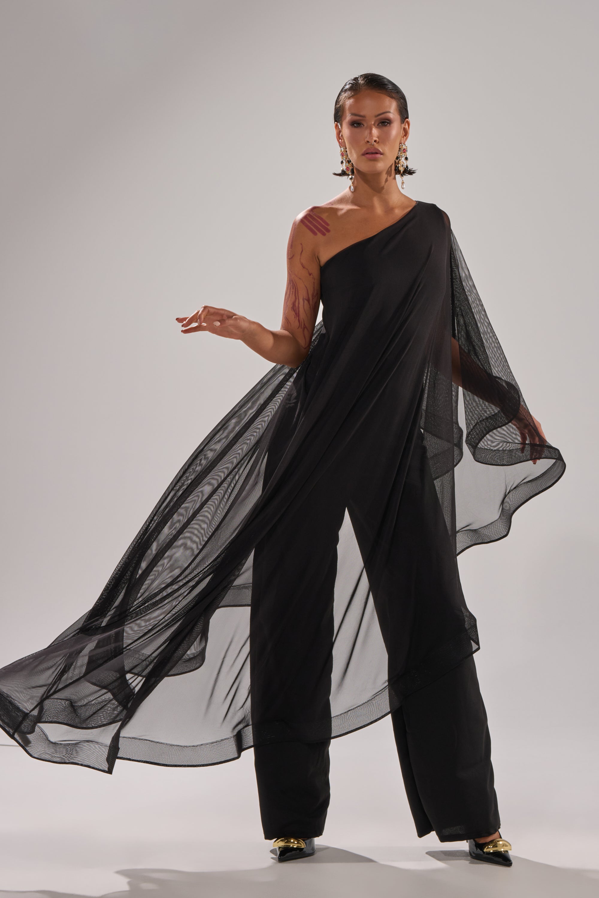 A person poses confidently in the SANDO MESH JUMPSUIT IN BLACK, a stylish one-shoulder design with sheer mesh and flowing wide-leg pants, accessorized with gold earrings and black shoes against a plain light background.