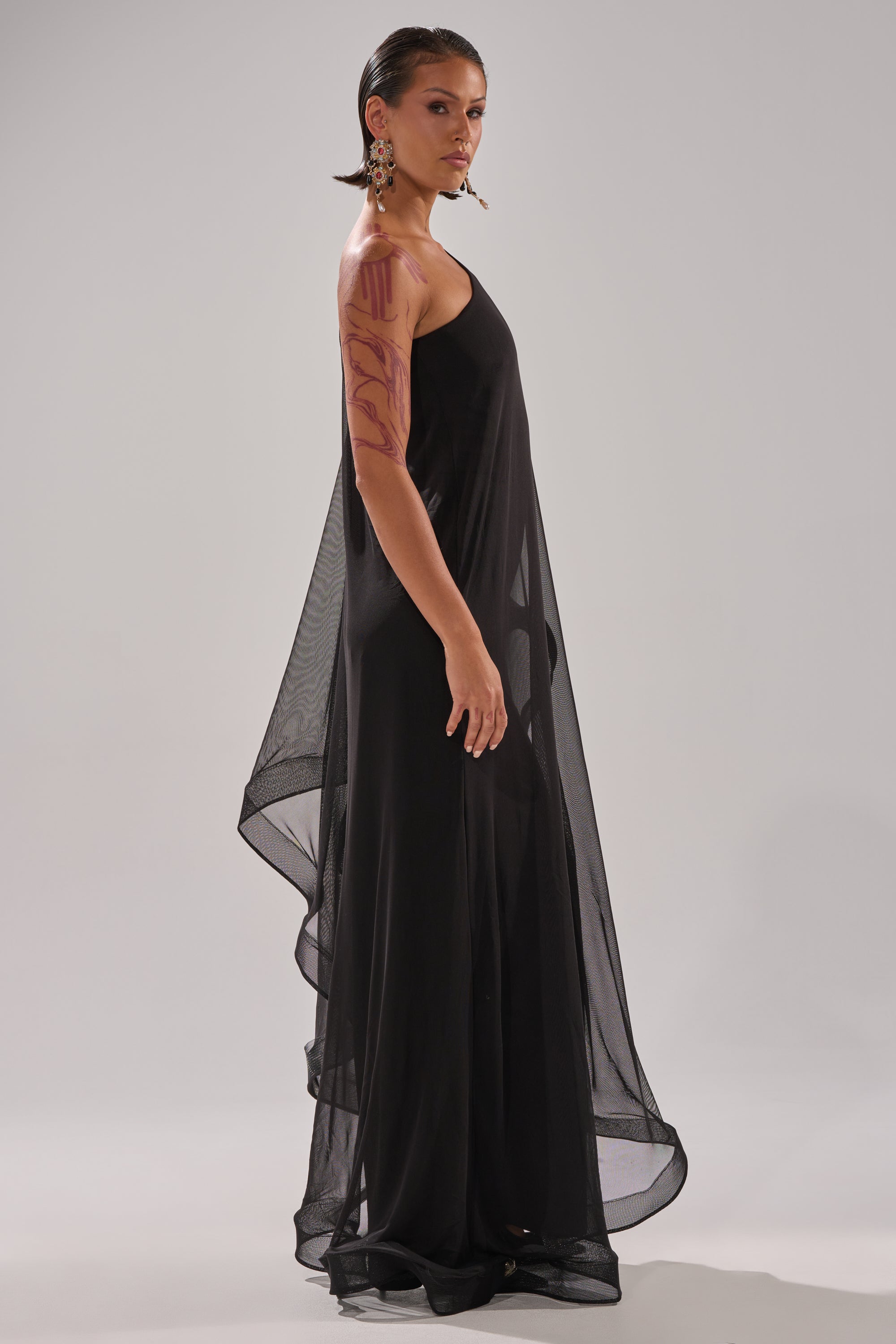 A person in profile wears the SANDO MESH JUMPSUIT IN BLACK, a flowing one-shoulder piece with sheer floor-length draping. They have slicked-back hair, large earrings, red arm markings, and stand against a plain light gray background.