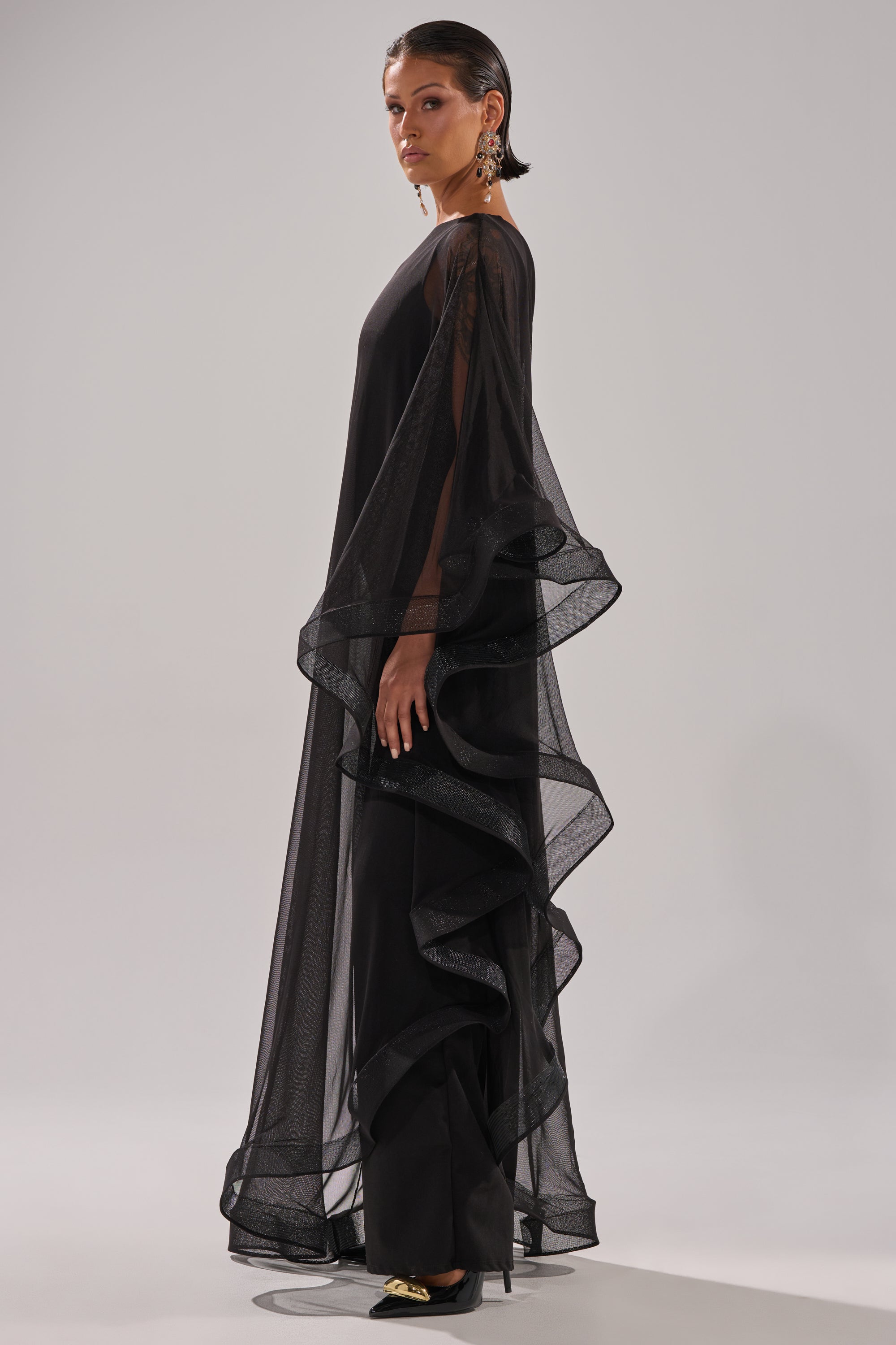 A woman stands in profile before a plain light background, wearing the SANDO MESH JUMPSUIT IN BLACK—dramatic and sheer with flowing ruffled layers. Slicked-back hair, statement earrings, and black pointed shoes with gold accents complete her look.