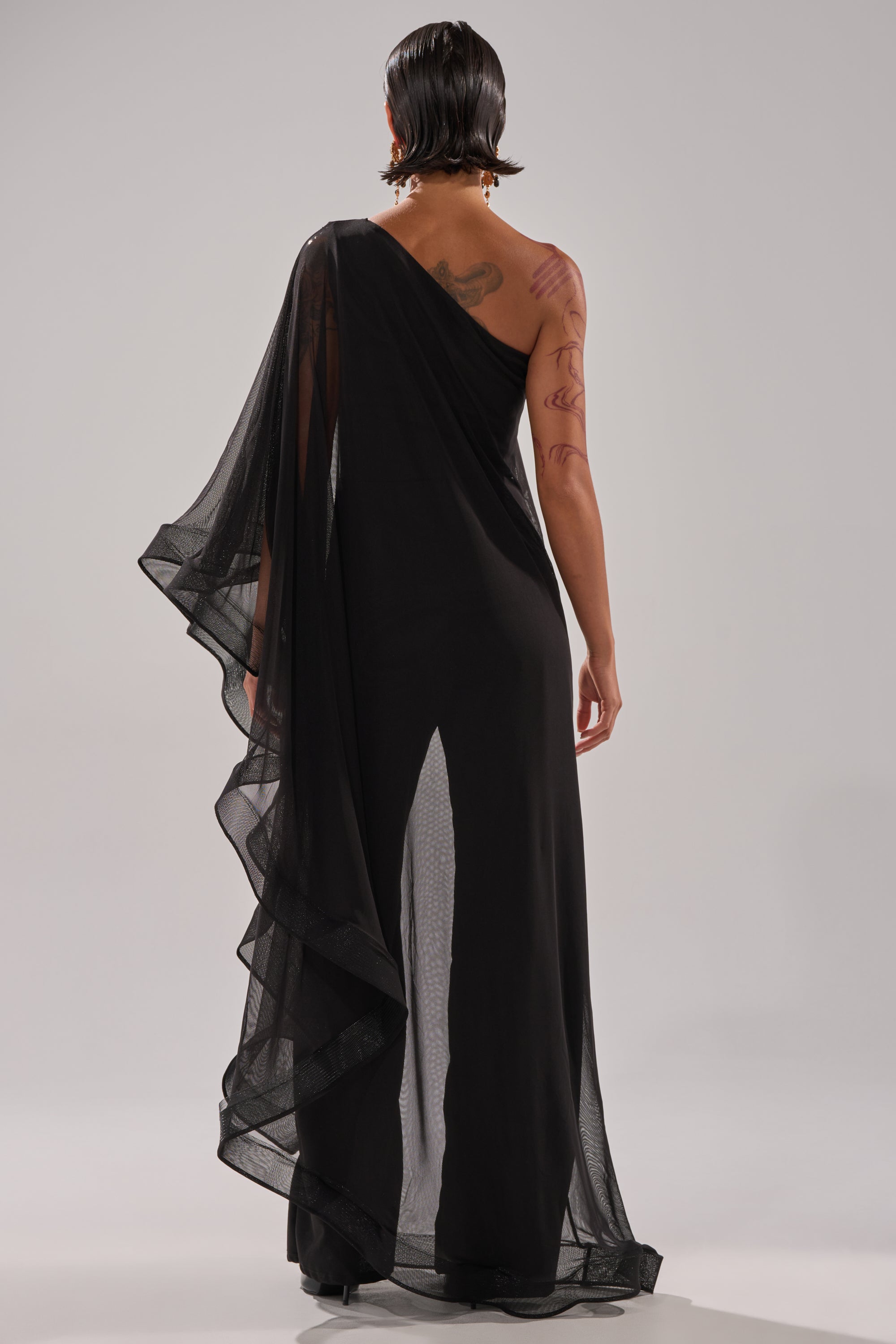 Wearing the SANDO MESH JUMPSUIT IN BLACK, a person with short, wet-look hair shows their back and arm tattoos through the long, one-shoulder sheer mesh design as they face away from the camera.