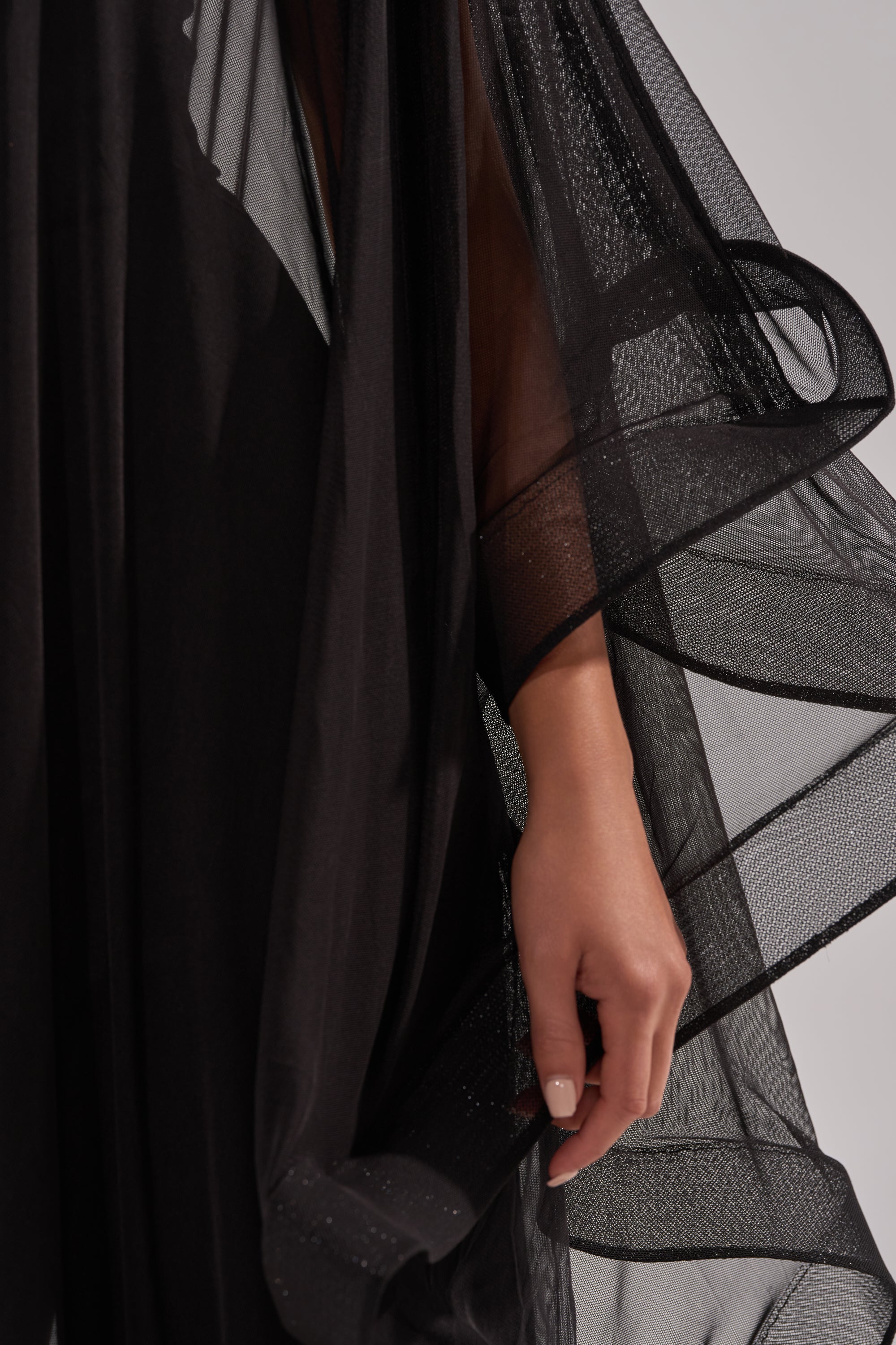 A close-up shows a relaxed hand holding the flowing edge of the SANDO MESH JUMPSUIT IN BLACK, highlighting its delicate, semi-transparent fabric against a neutral background.