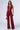 A woman stands modeling the LIVING MY WAY BURGUNDY JUMPSUIT, showcasing its super stretchy mesh fabric, deep V-neckline, and wide-leg pants. She is holding a small, round gold handbag in one hand and wears beige platform shoes. The background is plain and well-lit.