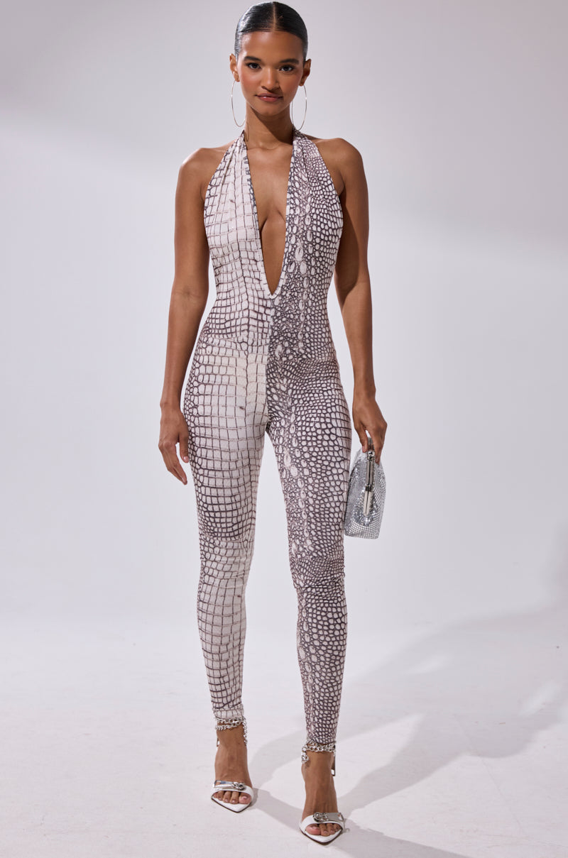 A woman stands confidently in the ON THE MOVE CROC PRINT JUMPSUIT, featuring a sleeveless deep V-neck halter design with a light brown croc pattern. She accessorizes with hoop earrings, strappy heels, a silver purse, and slicked-back hair.