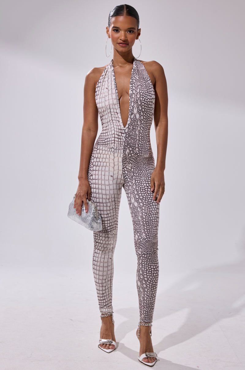 A woman poses against a plain light background wearing the ON THE MOVE CROC PRINT JUMPSUIT—sleeveless with a deep V-neck—paired with silver heels, large hoop earrings, and a silver clutch.