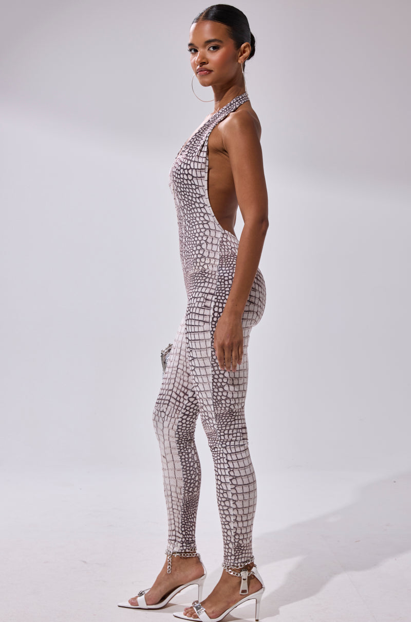 Woman wearing the ON THE MOVE CROC PRINT JUMPSUIT, sleeveless and backless, pairs it with white high-heeled sandals, standing sideways and looking at the camera against a plain white background.