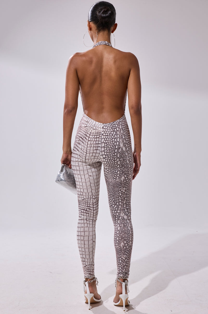 A woman faces away, wearing the ON THE MOVE CROC PRINT JUMPSUIT with a halter neckline, white high-heeled sandals, and a small silver handbag. Her hair is styled in a sleek bun.