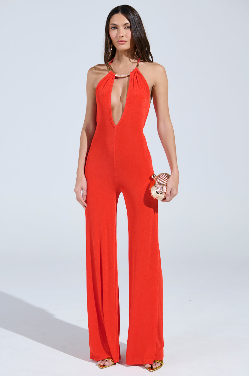 A woman confidently poses against a plain backdrop in the vibrant BACK IN TIME JUMPSUIT WITH RUCHING, featuring a deep neckline. She accessorizes with a small round purse and open-toed heels, completing the perfect ensemble for a night of dancing.