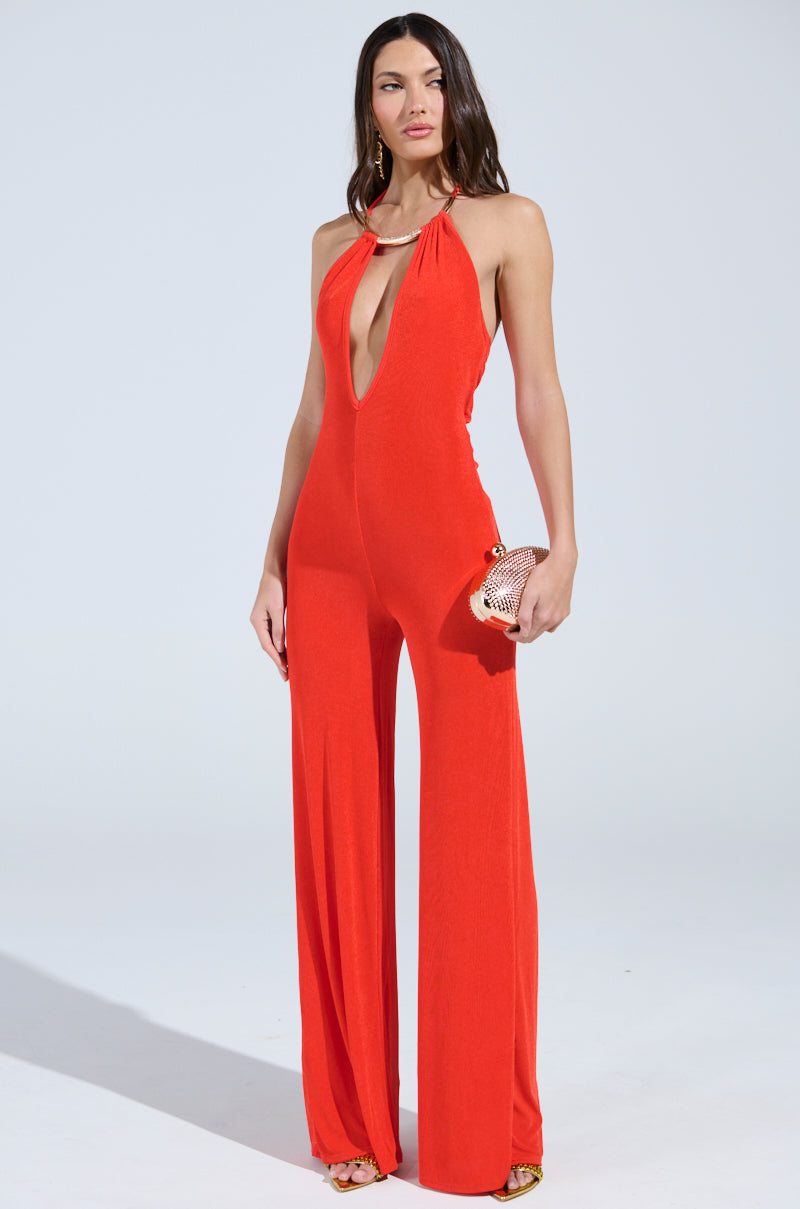 A confident person wears the BACK IN TIME JUMPSUIT WITH RUCHING, a sleeveless bright red disco outfit with a deep neckline. They accessorize with a small clutch and stylish heels, exuding flair against a plain light backdrop.