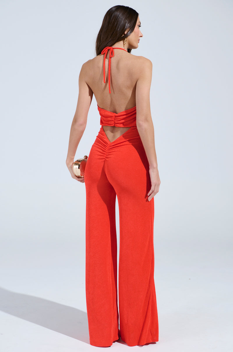 A woman poses against a plain background in the vibrant red BACK IN TIME JUMPSUIT WITH RUCHING, featuring an open back and wide-legged pants. A small clutch accents her ensemble, with her long hair cascading elegantly down her back.