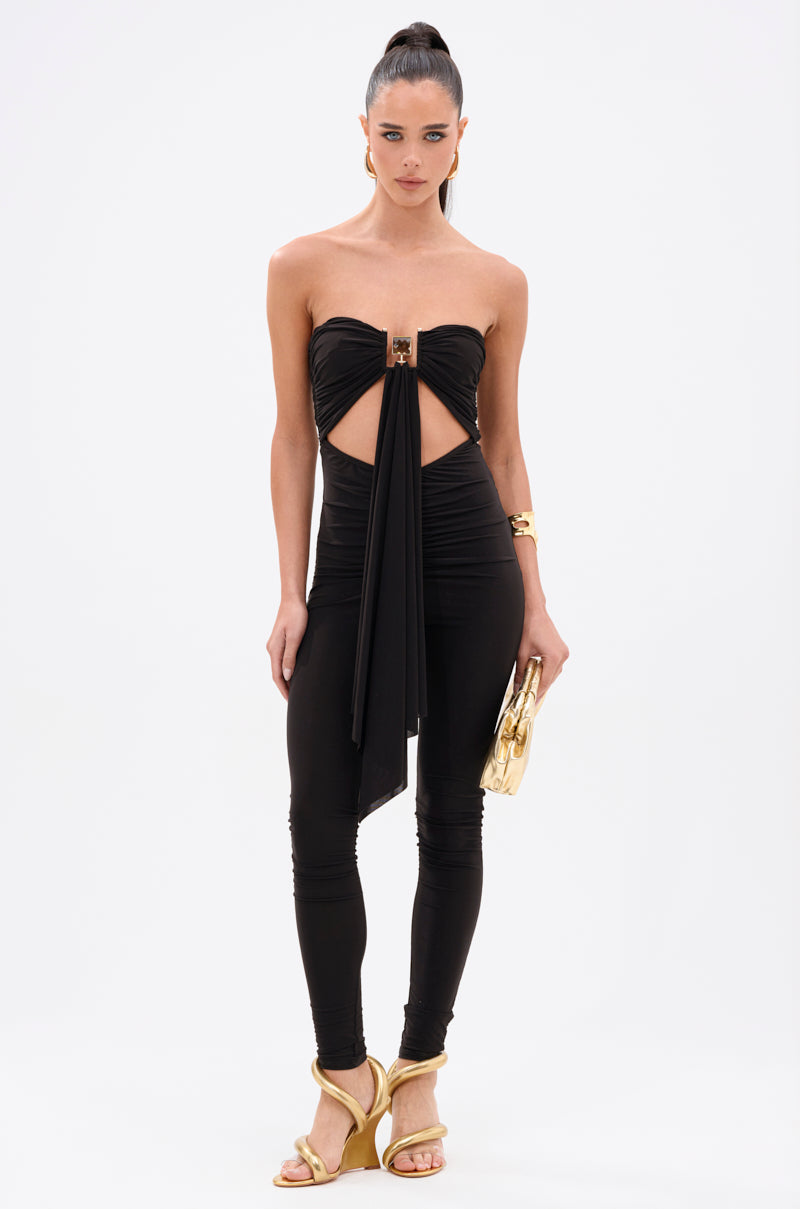 A woman wears the ONE OF A KIND CUTOUT JUMPSUIT in luxe stretch fabric with a strapless cutout, ruched detail, and draped accents, styled with a gold clutch, gold heeled sandals, a chic gold bracelet, and a high ponytail.