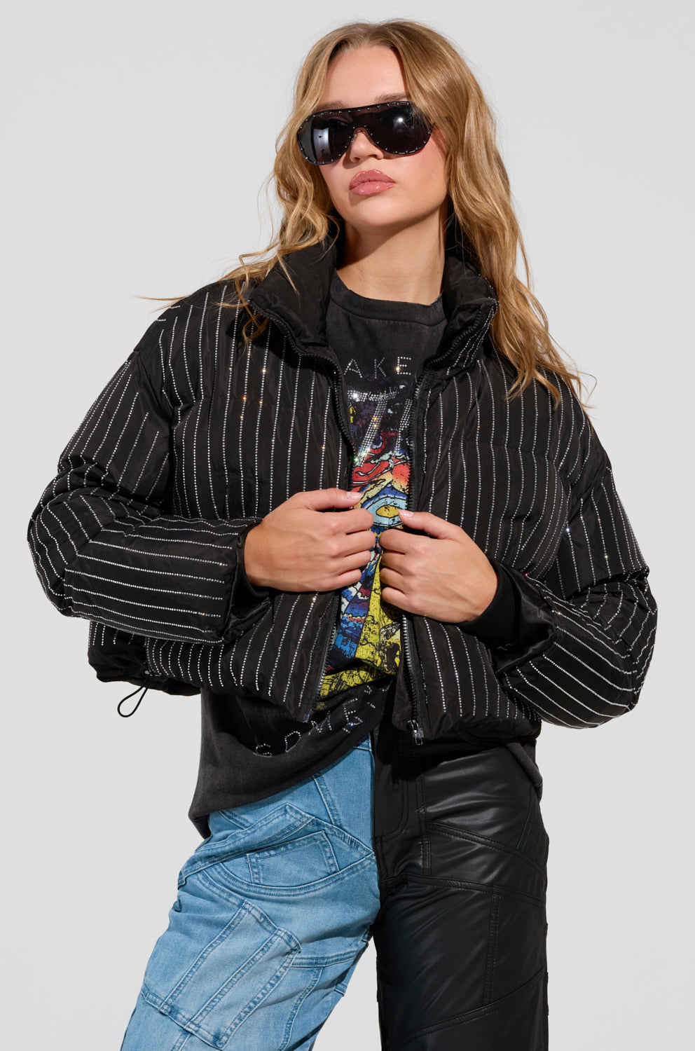 A person with long hair wears sunglasses, the COME THIS WAY EMBELLISHED PUFFER COAT, and a graphic t-shirt. They pair blue denim with sleek black leather pants, standing confidently against a plain background.