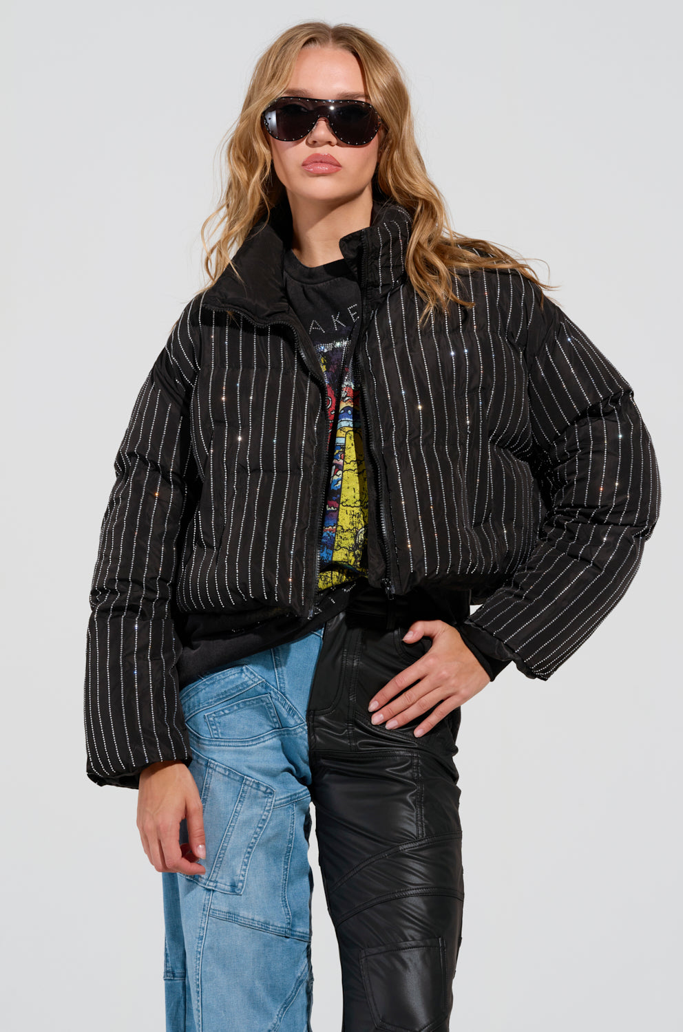 A person with long hair and sunglasses poses confidently, wearing the "COME THIS WAY EMBELLISHED PUFFER COAT" over a graphic t-shirt. They have mismatched pants—one leg in blue denim and the other in black, standing against a plain background.
