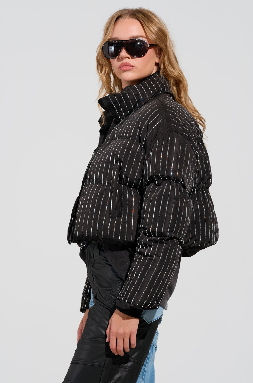 A person clad in the COME THIS WAY EMBELLISHED PUFFER COAT and sunglasses poses confidently. Their long, wavy hair cascades over the quilted design of the jacket, set against a plain light gray background.