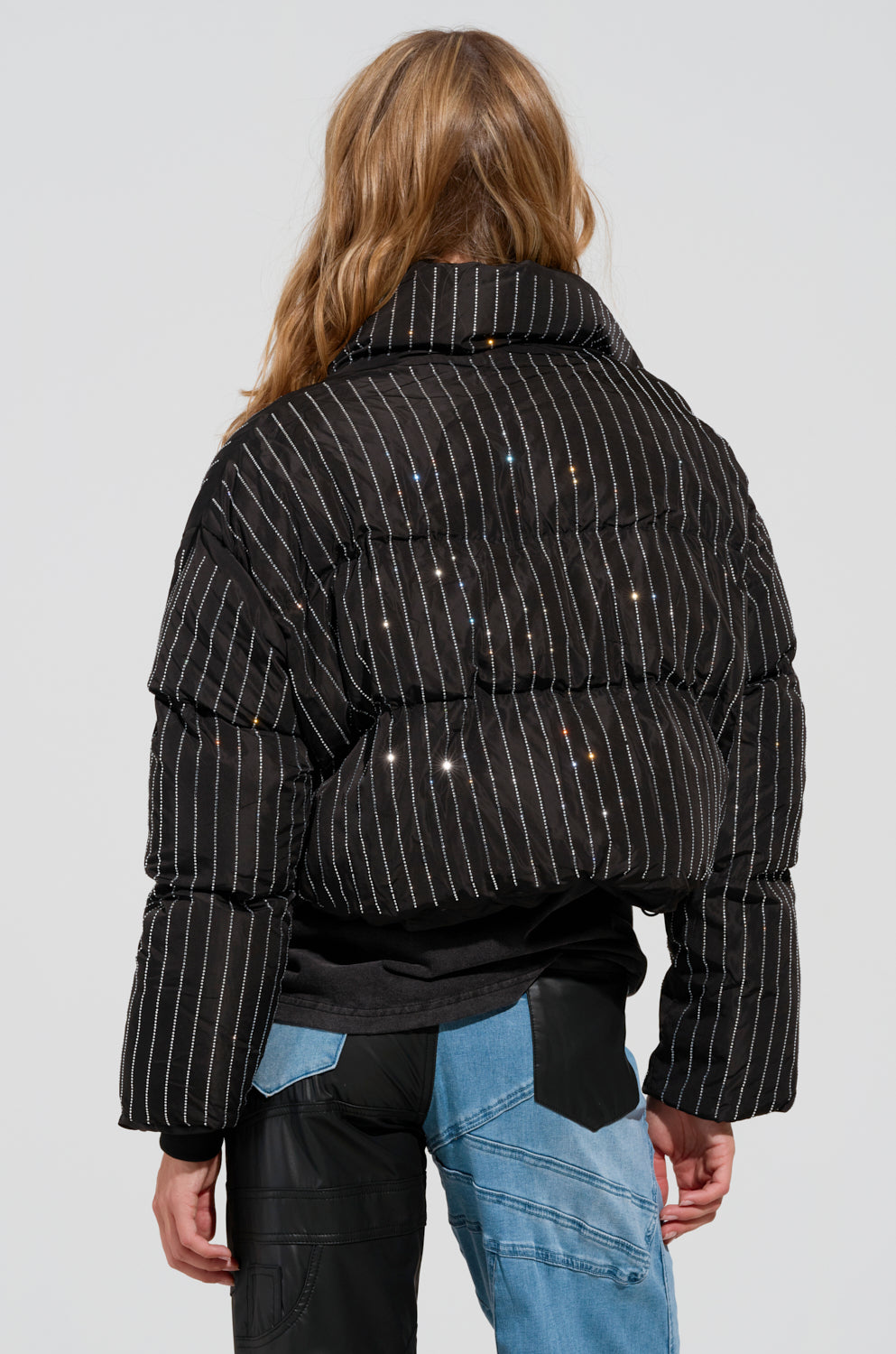 Dressed in the COME THIS WAY EMBELLISHED PUFFER COAT, adorned with white vertical stripes and delicate light reflections, a long-haired individual stands with their back to the camera. Their pants are designed with a black and blue patchwork pattern, enhancing the striking quilted aesthetics of their outfit.