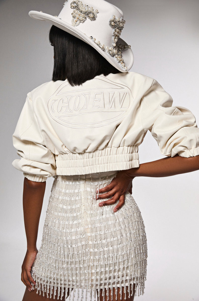 A woman stands with her back turned, wearing the INTO YOU CROPPED MOTO jacket featuring an embossed logo. She pairs it with a white beaded fringed skirt and an embellished wide-brimmed hat, posing with hands on hips against a neutral backdrop.