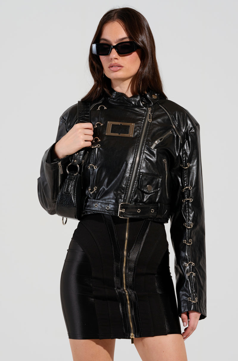 A woman wearing the WHY SO SERIOUS METAL TRIM MOTO jacket, which features stylish metallic hardware detailing, paired with a matching black skirt. She has long dark hair and large black sunglasses, perfectly capturing biker-chic outerwear vibes. Her confident pose stands out against a plain background.