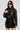 A woman wearing the WHY SO SERIOUS METAL TRIM MOTO jacket, which features stylish metallic hardware detailing, paired with a matching black skirt. She has long dark hair and large black sunglasses, perfectly capturing biker-chic outerwear vibes. Her confident pose stands out against a plain background.