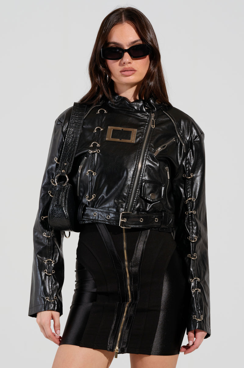 Dressed in the WHY SO SERIOUS METAL TRIM MOTO jacket, a woman exudes biker-chic style with its shiny black faux leather and metallic hardware detailing. She effortlessly pairs it with a black zippered dress, her long hair flowing and sunglasses on, while carrying a matching black handbag against the plain white backdrop.
