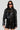 Dressed in the WHY SO SERIOUS METAL TRIM MOTO jacket, a woman exudes biker-chic style with its shiny black faux leather and metallic hardware detailing. She effortlessly pairs it with a black zippered dress, her long hair flowing and sunglasses on, while carrying a matching black handbag against the plain white backdrop.