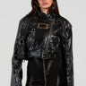 Dressed in the WHY SO SERIOUS METAL TRIM MOTO jacket, a woman exudes biker-chic style with its shiny black faux leather and metallic hardware detailing. She effortlessly pairs it with a black zippered dress, her long hair flowing and sunglasses on, while carrying a matching black handbag against the plain white backdrop.