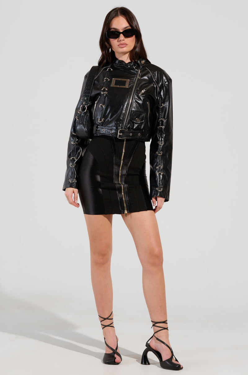 A woman exudes confidence, sporting sleek biker-chic outerwear: the WHY SO SERIOUS METAL TRIM MOTO, paired with sunglasses and a black dress featuring a front zipper. Her look is completed by strappy black heels against a plain backdrop.