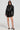 A woman exudes confidence, sporting sleek biker-chic outerwear: the WHY SO SERIOUS METAL TRIM MOTO, paired with sunglasses and a black dress featuring a front zipper. Her look is completed by strappy black heels against a plain backdrop.