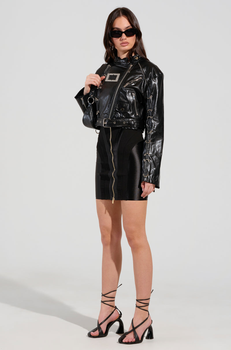 A woman radiates confidence as she poses in the WHY SO SERIOUS METAL TRIM MOTO jacket, which features sleek metallic hardware, paired with a fitted black skirt adorned with a front zipper. Her strappy black heels and dark sunglasses flawlessly complete her biker-chic look against a plain backdrop.