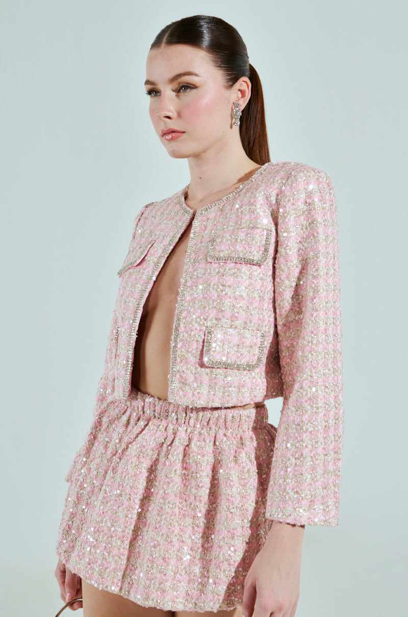With long hair in a sleek ponytail, they confidently wear the ALINNA EMBELLISHED TWEED BLAZER and a high-waisted sparkling pink textured skirt. The cropped silhouette features rhinestone trim as they pose against a light background with their earrings softly glinting.