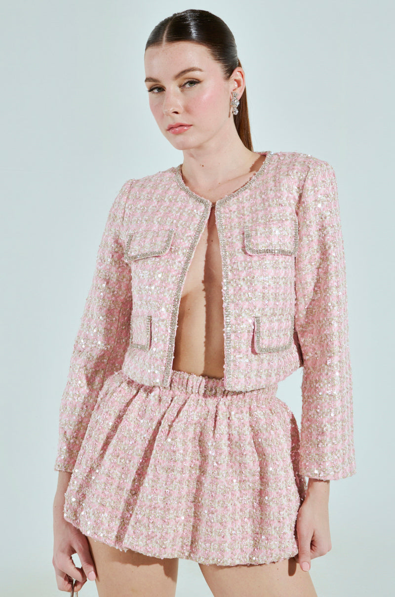 A woman models the ALINNA EMBELLISHED TWEED BLAZER, a chic cropped pink jacket paired elegantly with a high-waisted skirt. Her slicked-back hair and small hoop earrings accentuate the ensemble against a plain, light background.