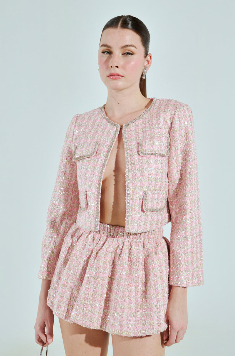 Against a plain backdrop, a person wears a pink textured two-piece outfit: the ALINNA EMBELLISHED TWEED BLAZER paired with a matching skirt. The open blazer reveals their style, their hair is tied back, and they hold an item in their left hand.