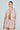 Against a plain backdrop, a person wears a pink textured two-piece outfit: the ALINNA EMBELLISHED TWEED BLAZER paired with a matching skirt. The open blazer reveals their style, their hair is tied back, and they hold an item in their left hand.
