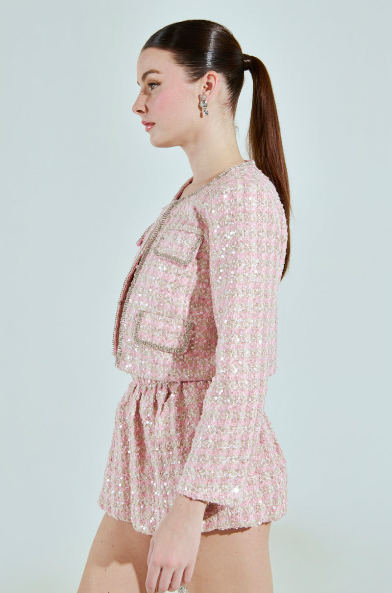A woman in the ALINNA EMBELLISHED TWEED BLAZER with rhinestone trim and matching shorts stands in profile against a plain background. Her hair is styled in a ponytail, and small hoop earrings perfectly complement her chic cropped silhouette.