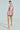 A woman showcases the ALINNA EMBELLISHED TWEED BLAZER, a short pink jacket with two large front pockets, paired with a matching skirt and fluffy pink heels. Her sleek hair complements the ensemble's elegant cropped silhouette against a plain white background.