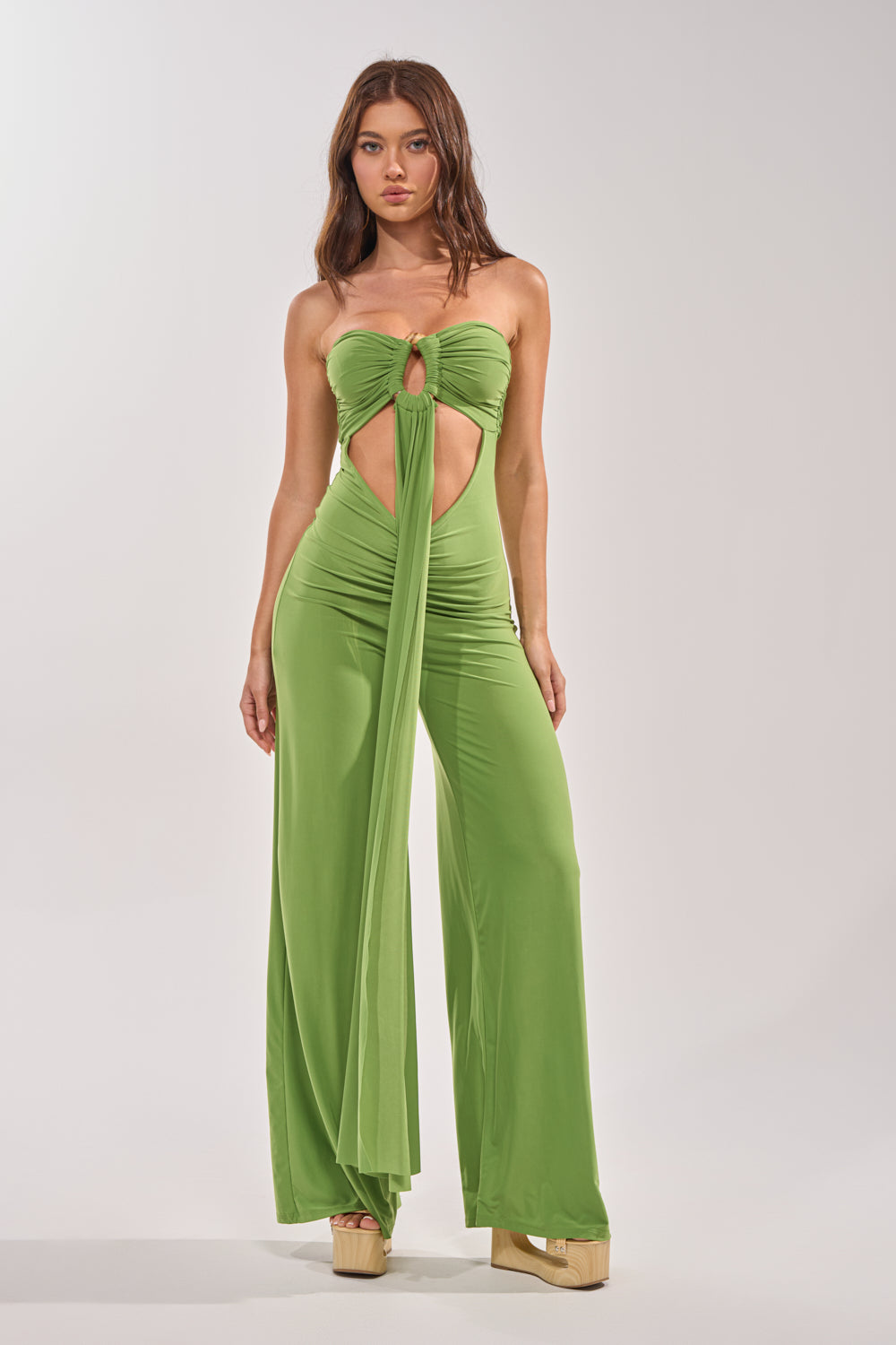 A woman faces forward in the PICK ME UP STRAPLESS JUMPSUIT IN GREEN featuring a strapless neckline, wide-leg silhouette, keyhole cutout, and long sash. She wears beige platform sandals. The background is plain and light-colored. 2