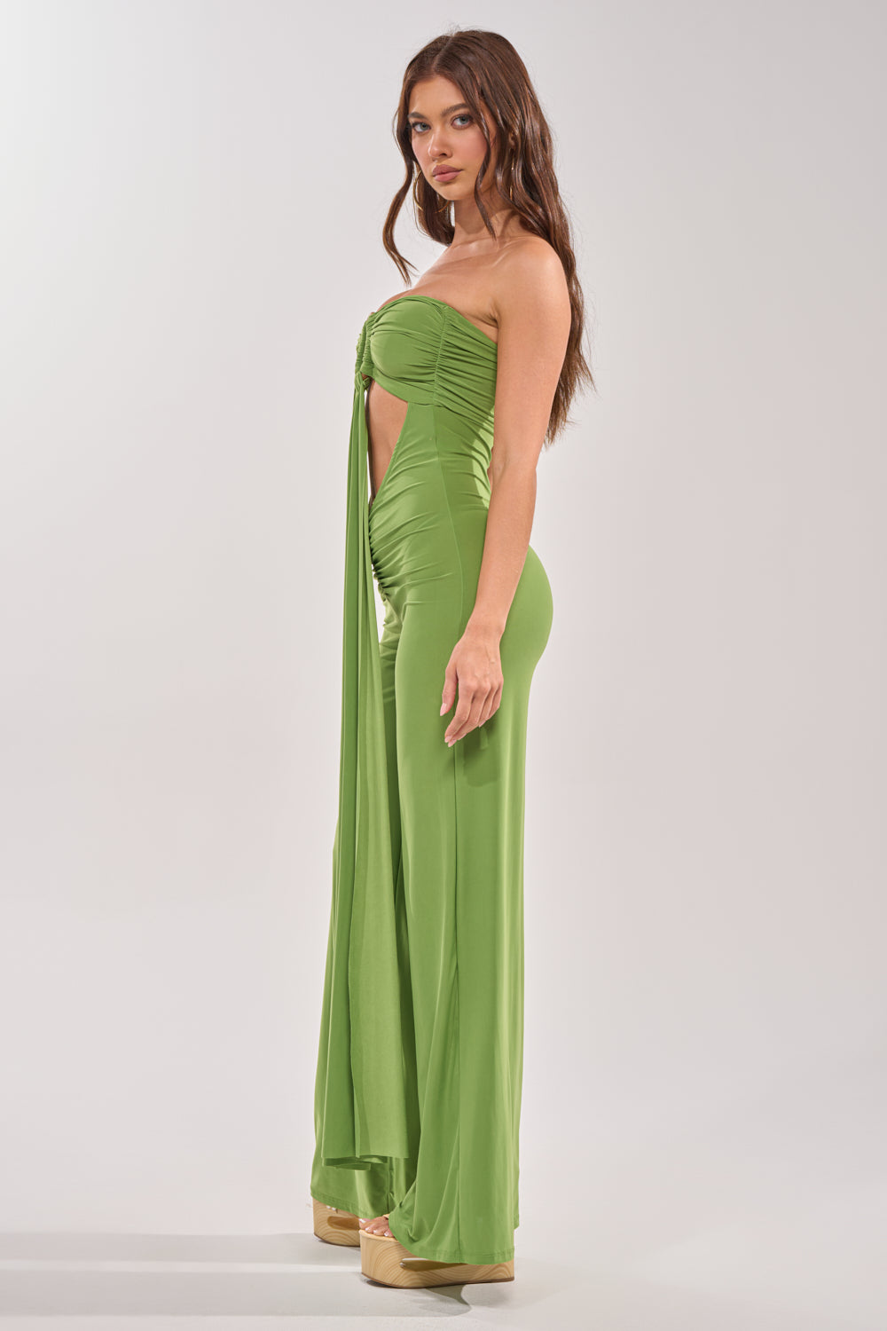 A woman models the PICK ME UP STRAPLESS JUMPSUIT IN GREEN, featuring a strapless design, front cutout, and wide-leg silhouette. She stands sideways in platform shoes with long brown hair—ideal for chic resort styling. 3