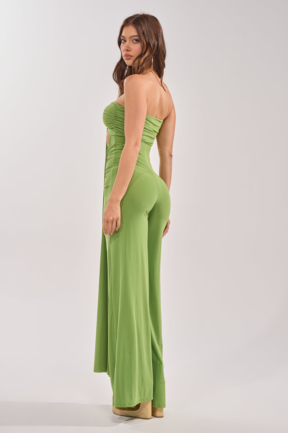 A woman models the PICK ME UP STRAPLESS JUMPSUIT IN GREEN with a wide-leg silhouette, paired with platform heels. She stands side-on and looks over her shoulder against a plain light background. 4