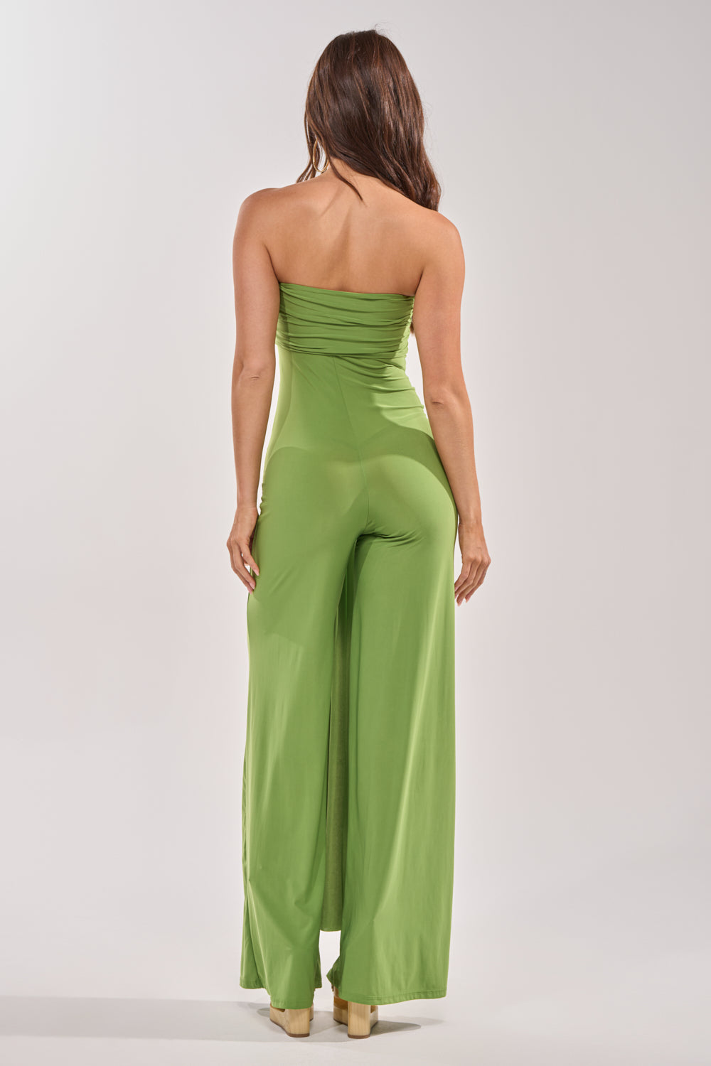 A woman with long brown hair faces away, wearing the PICK ME UP STRAPLESS JUMPSUIT IN GREEN featuring a wide-leg silhouette and beige platform heels against a plain light background. 5
