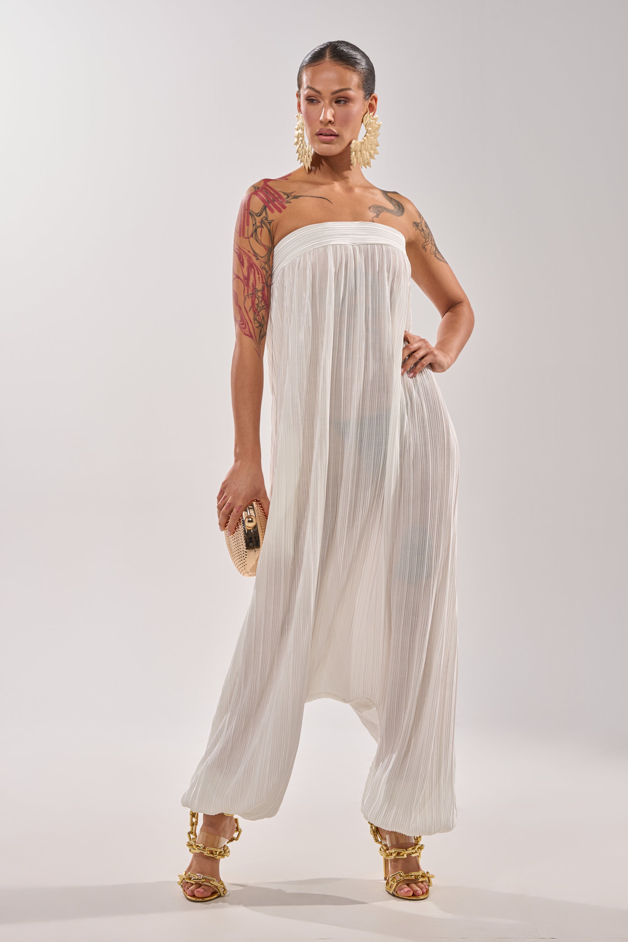 A woman models the FROM THE CITY DROP CROTCH JUMPSUIT IN WHITE with flowing pleats, paired with gold sandals, large gold earrings, a textured clutch, and visible arm tattoos. Her hair is pulled back as she stands against a plain background. 0