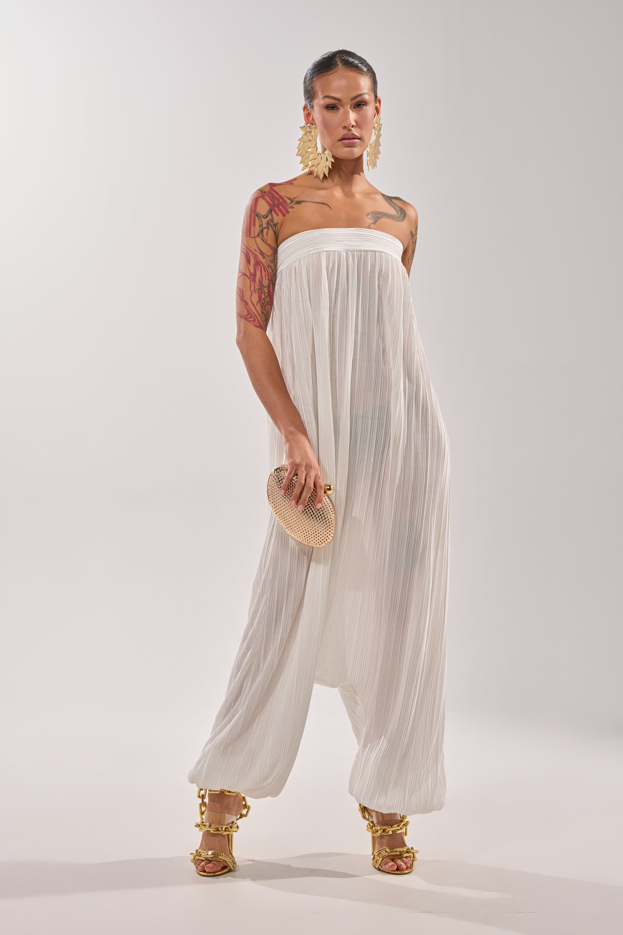 A woman poses confidently in the FROM THE CITY DROP CROTCH JUMPSUIT IN WHITE, accessorized with large gold earrings, gold heels, a small textured clutch, and a bold arm tattoo against a plain light background. 1