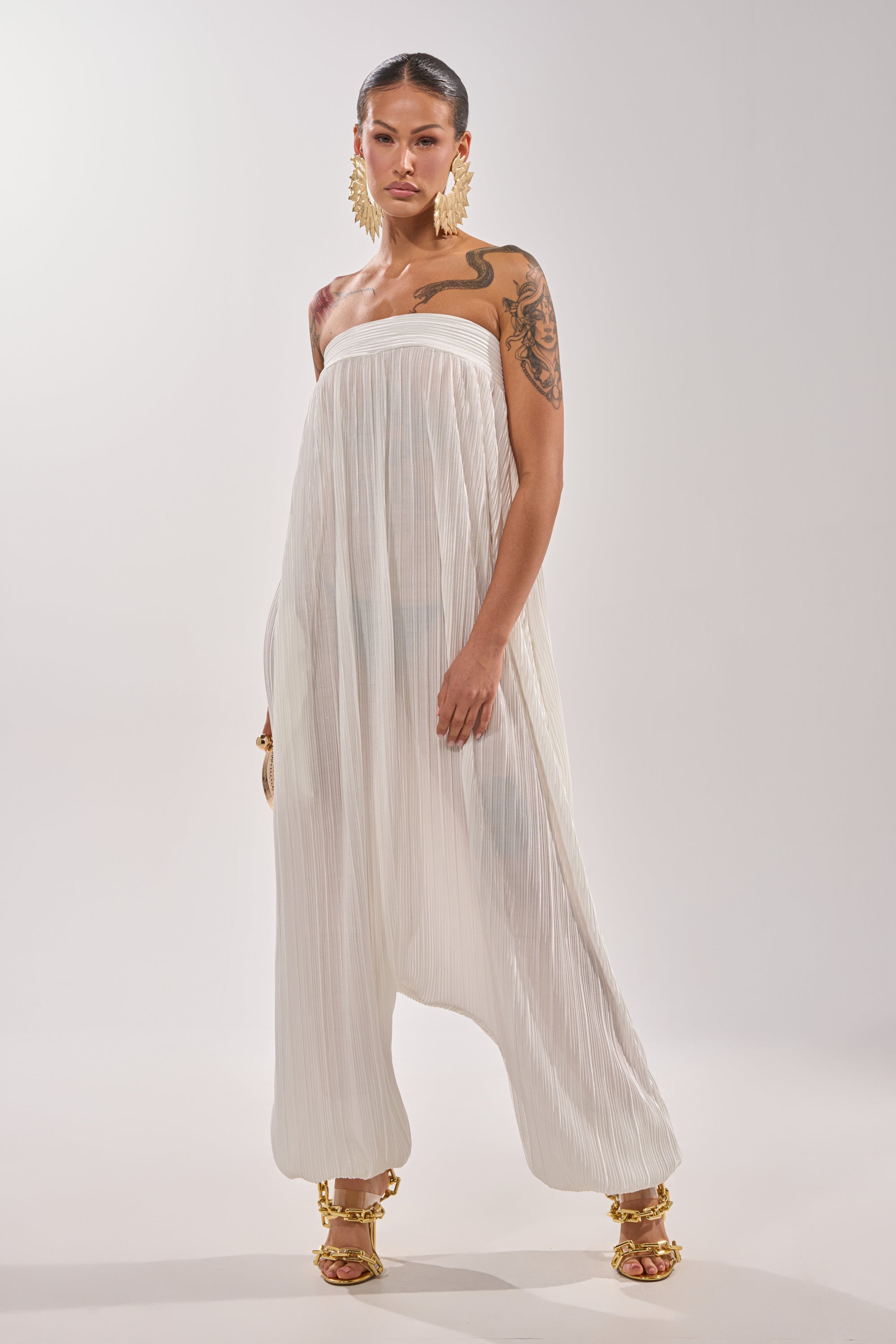 A woman poses confidently in the FROM THE CITY DROP CROTCH JUMPSUIT IN WHITE, featuring a strapless design and loose, wide legs. She accessorizes with gold earrings, gold heels, and displays upper arm tattoos against a light background. 2
