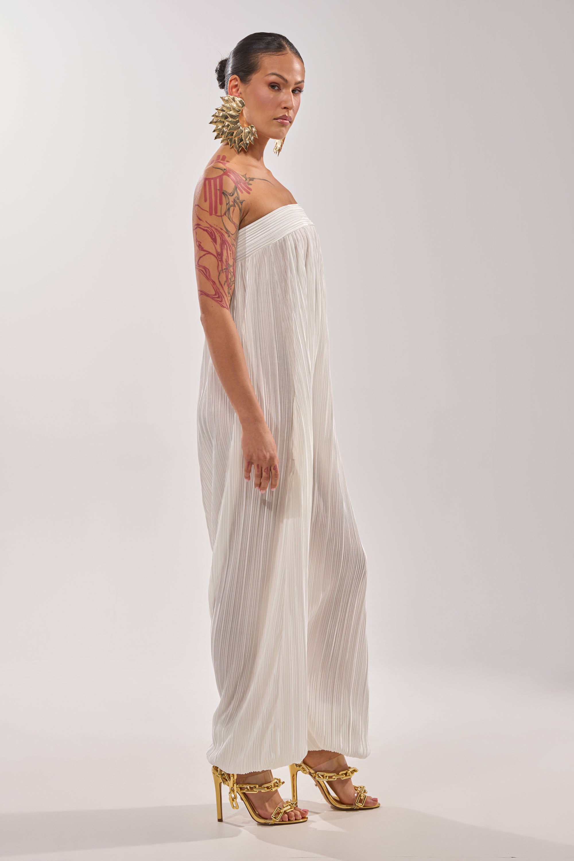 A woman with a tattooed arm stands sideways against a plain background, wearing the FROM THE CITY DROP CROTCH JUMPSUIT IN WHITE with gold high-heeled sandals and statement earrings. Her hair is styled in a neat bun. 3