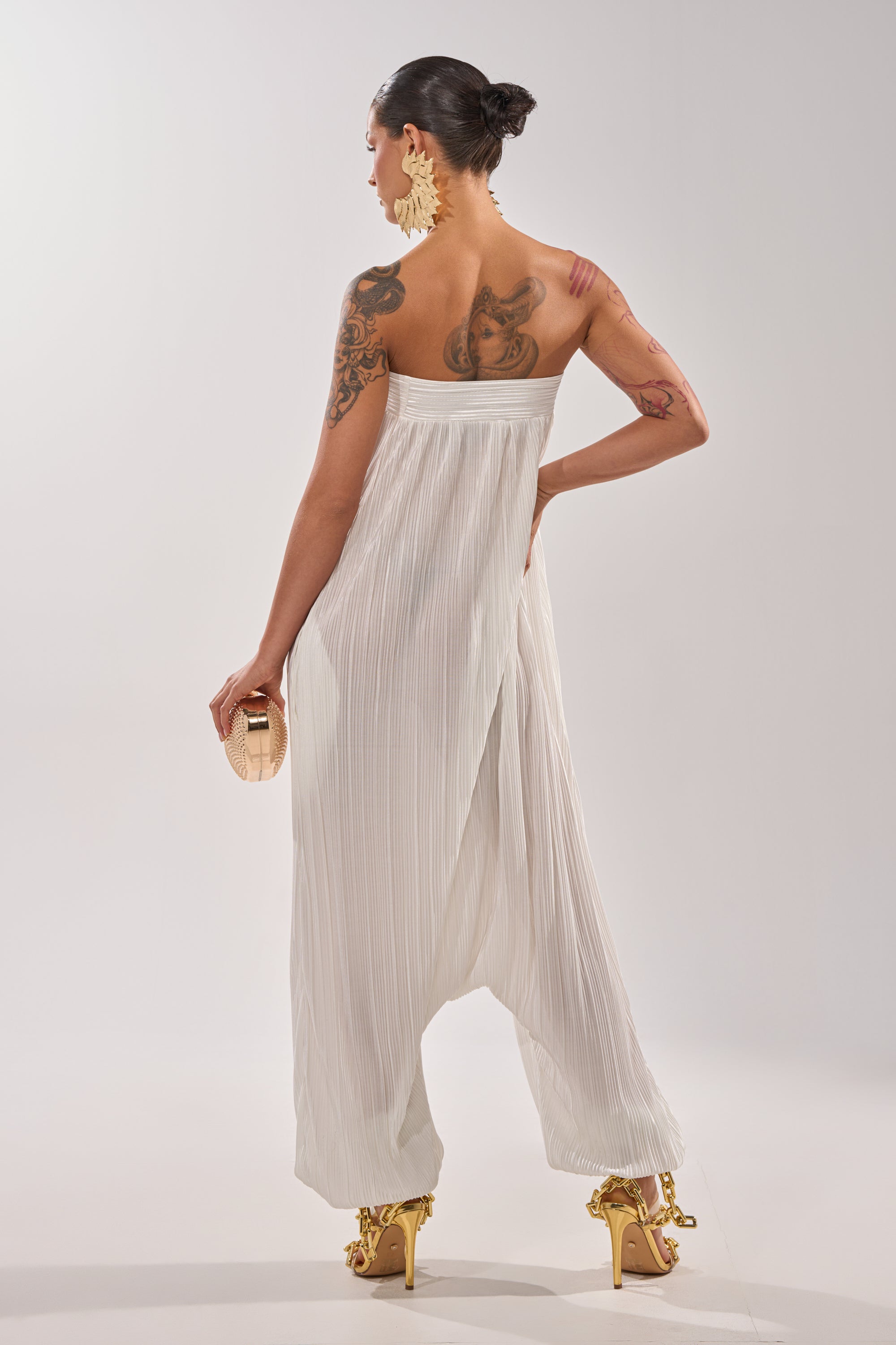 A tattooed woman faces away in the FROM THE CITY DROP CROTCH JUMPSUIT IN WHITE, paired with gold high heels, a beige clutch, and bold statement earrings. 4