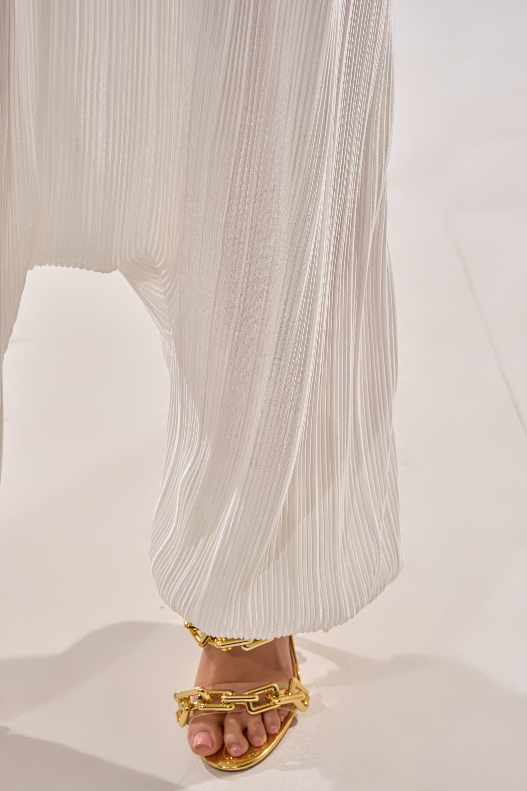 A close-up of a person in the FROM THE CITY DROP CROTCH JUMPSUIT IN WHITE, pleated and strapless, paired with gold chain sandals—the hem gently grazes the ground for an effortlessly chic look. 6