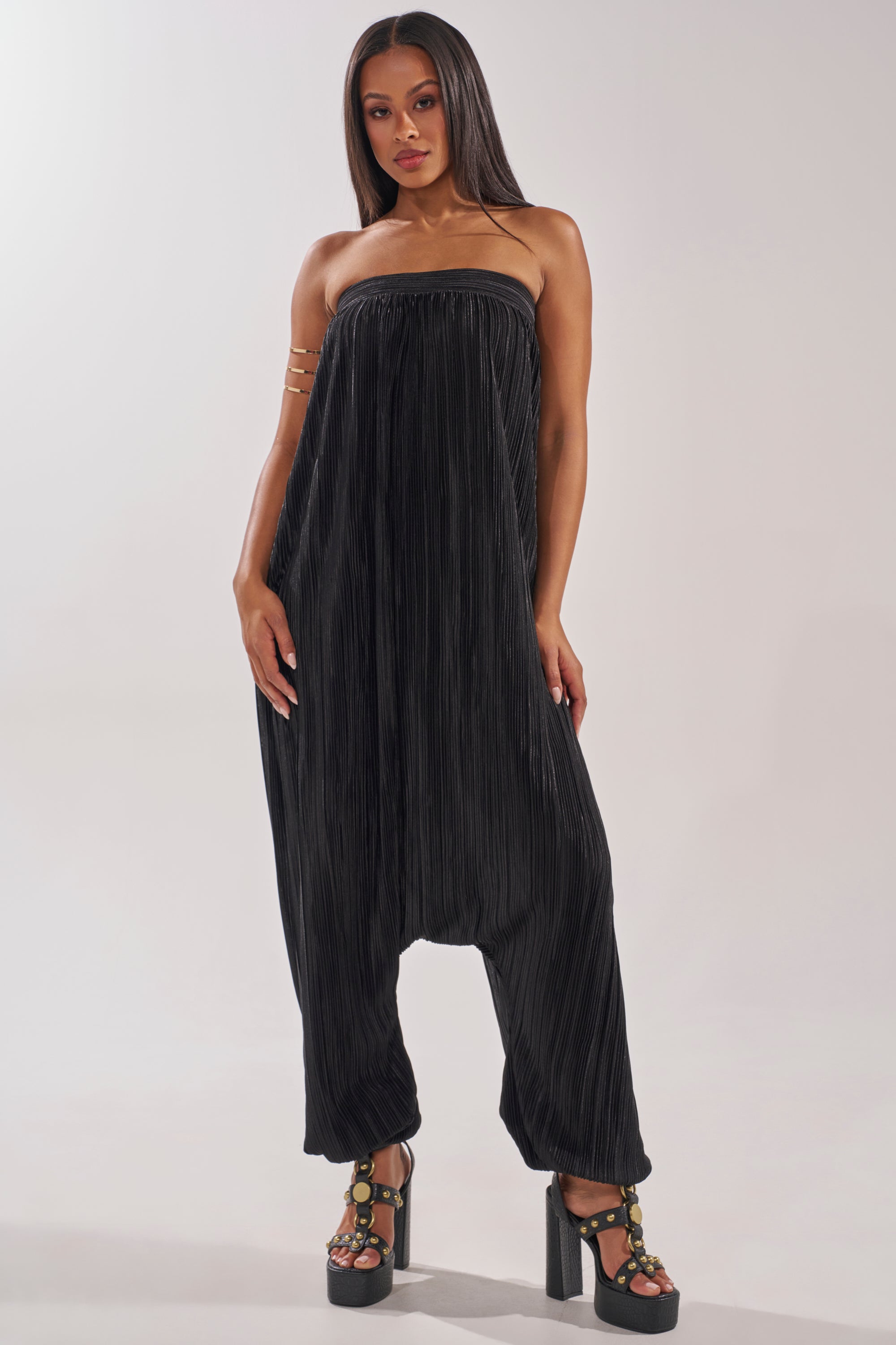 A woman faces forward in the FROM THE CITY DROP CROTCH JUMPSUIT IN BLACK, a loose, pleated, strapless evening resort style. She pairs it with black platform sandals featuring gold accents, her long straight hair worn down and expression neutral. 0