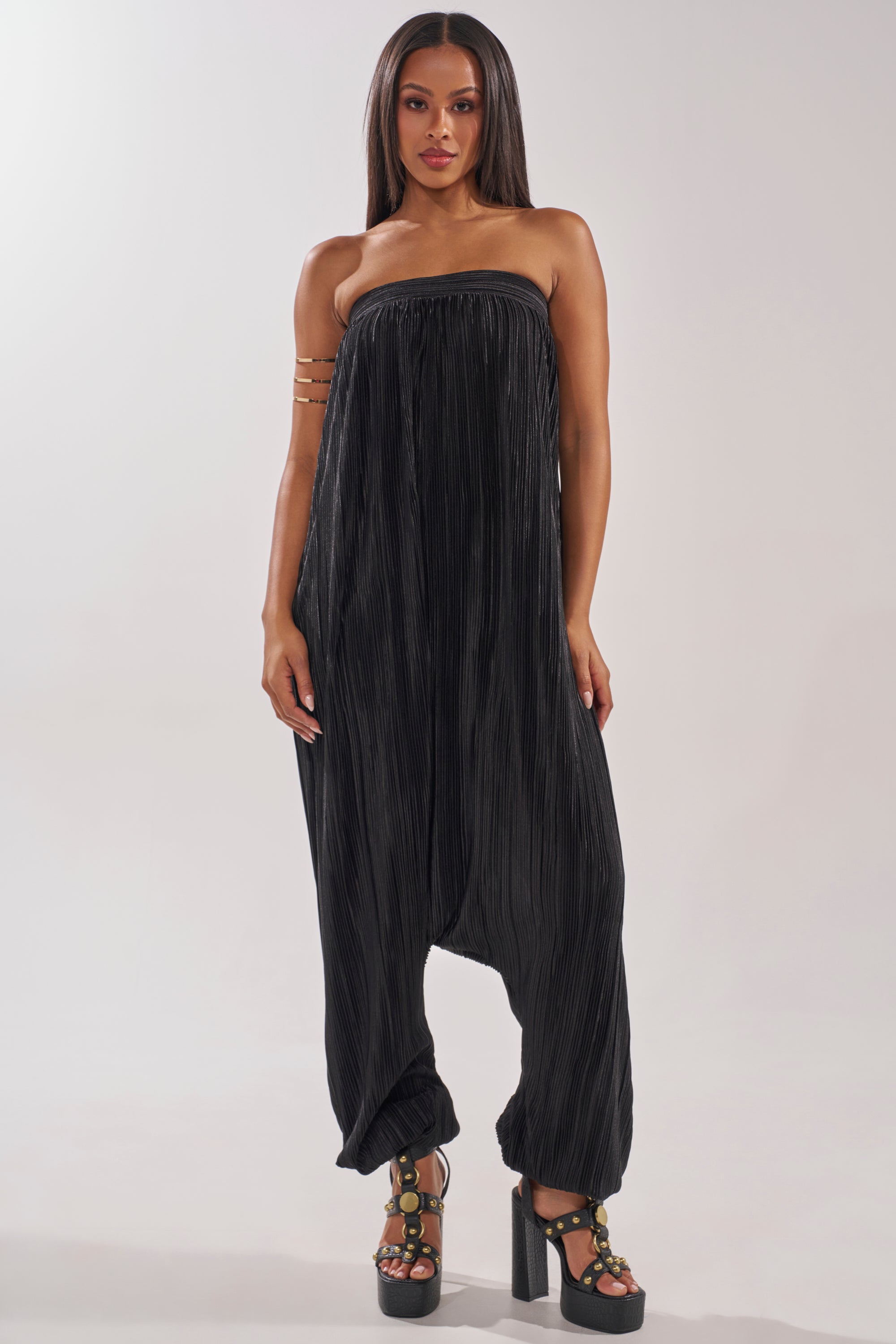 A woman stands against a plain background wearing the FROM THE CITY DROP CROTCH JUMPSUIT IN BLACK, featuring wide pleated legs gathered at the ankles, styled with black platform heels and an arm cuff—perfect for evening resort wear. 1