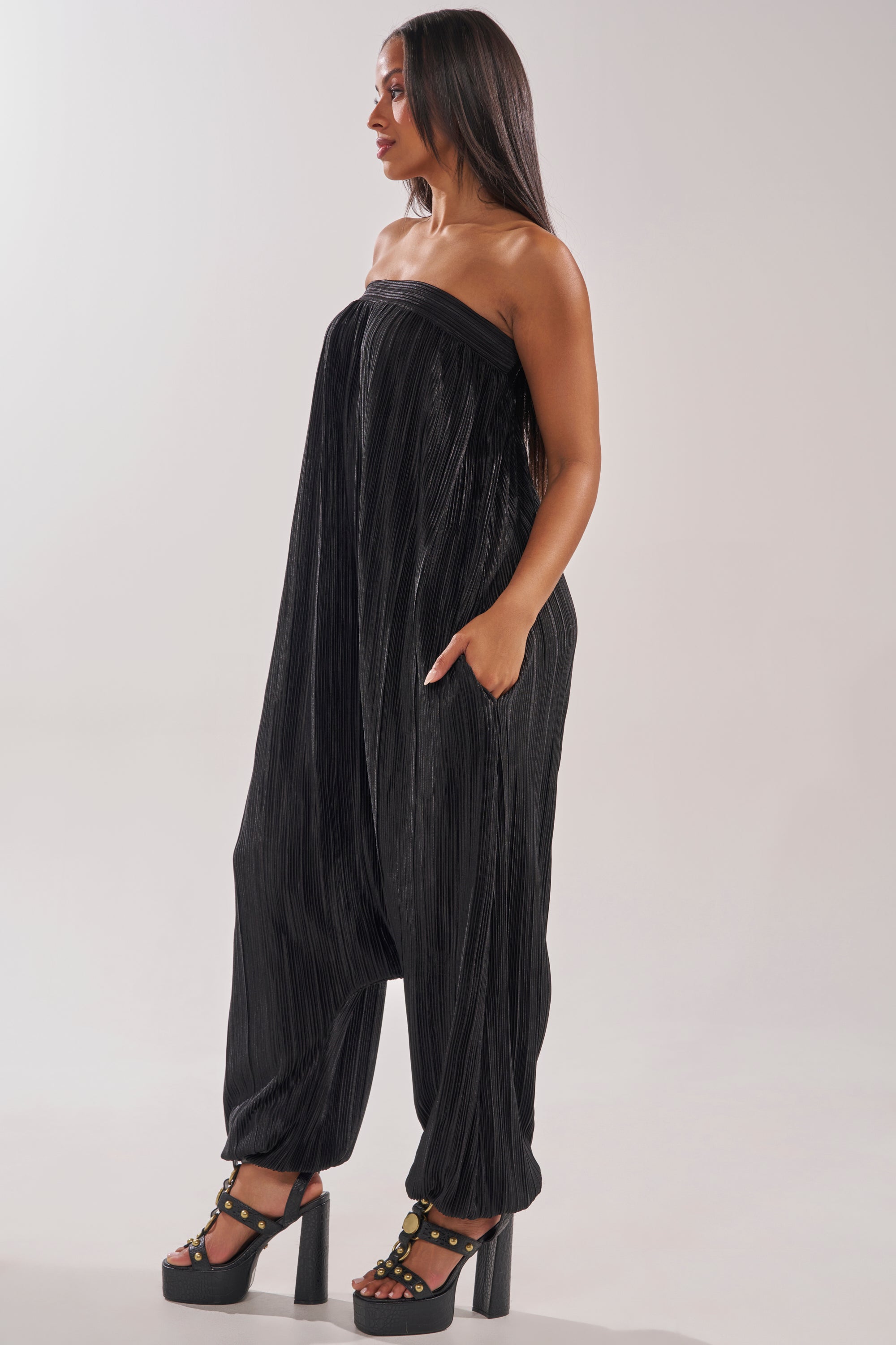 Woman in profile wears the FROM THE CITY DROP CROTCH JUMPSUIT IN BLACK, a strapless design with loose pleats and pockets, paired with high platform sandals—offering a chic, modern resort look against a plain light background. 2