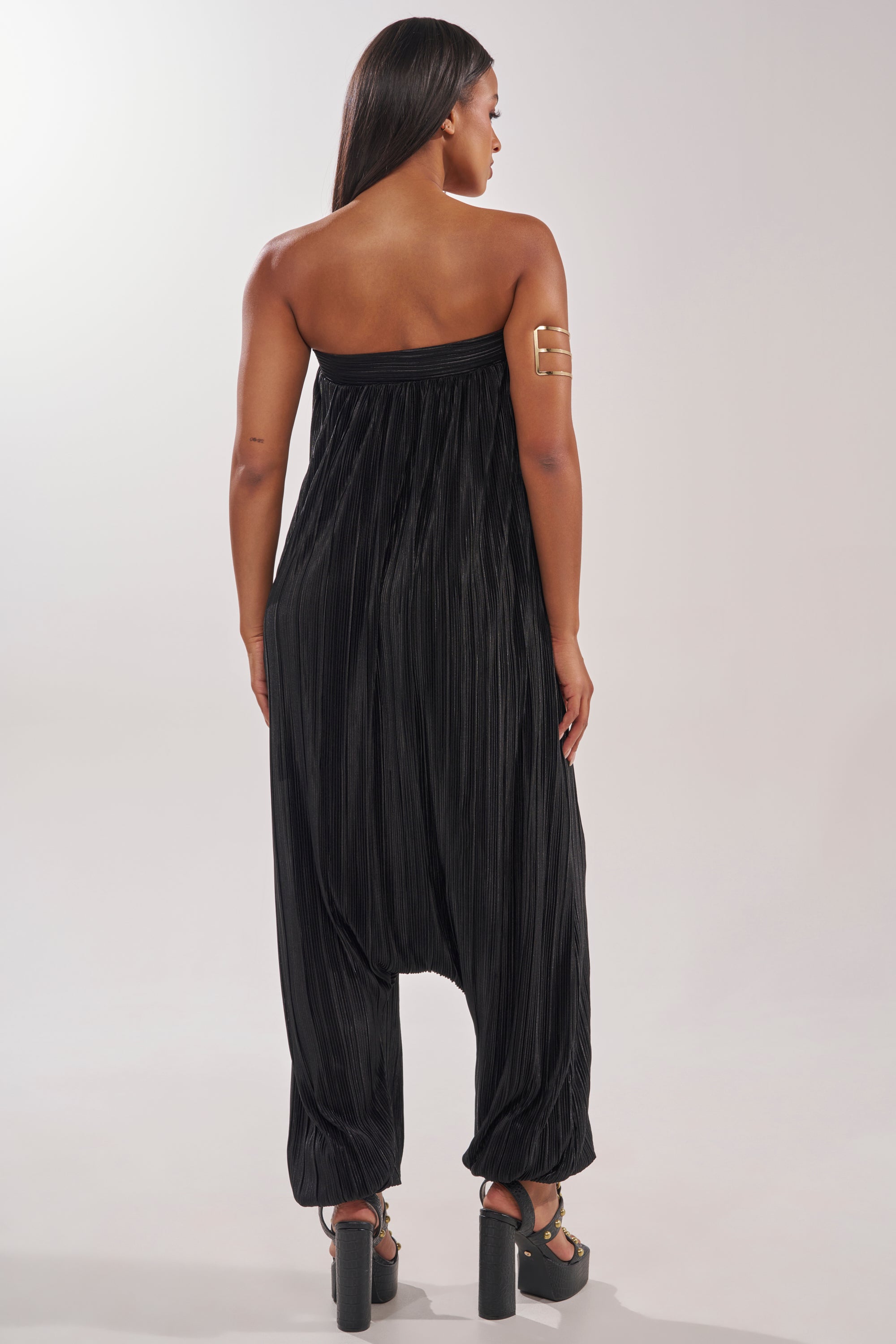 A woman with long straight hair models the FROM THE CITY DROP CROTCH JUMPSUIT IN BLACK, a strapless pleated style perfect for resort wear, paired with black platform sandals and a gold arm cuff against a light, plain background. 3