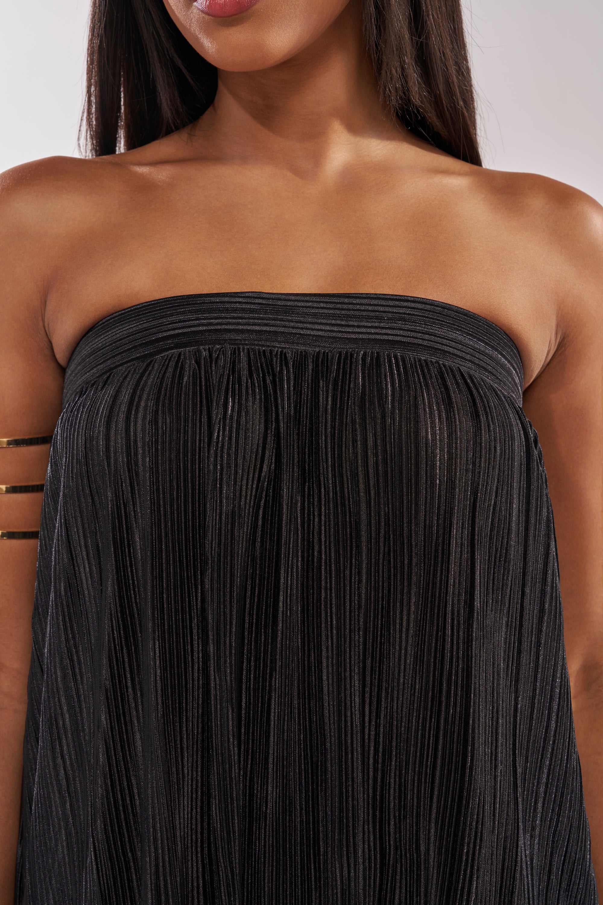 A woman wears the FROM THE CITY DROP CROTCH JUMPSUIT IN BLACK with pleated details and a metallic armband. The image shows her lower face, neck, shoulders, and upper chest—ideal for chic resort wear. 4