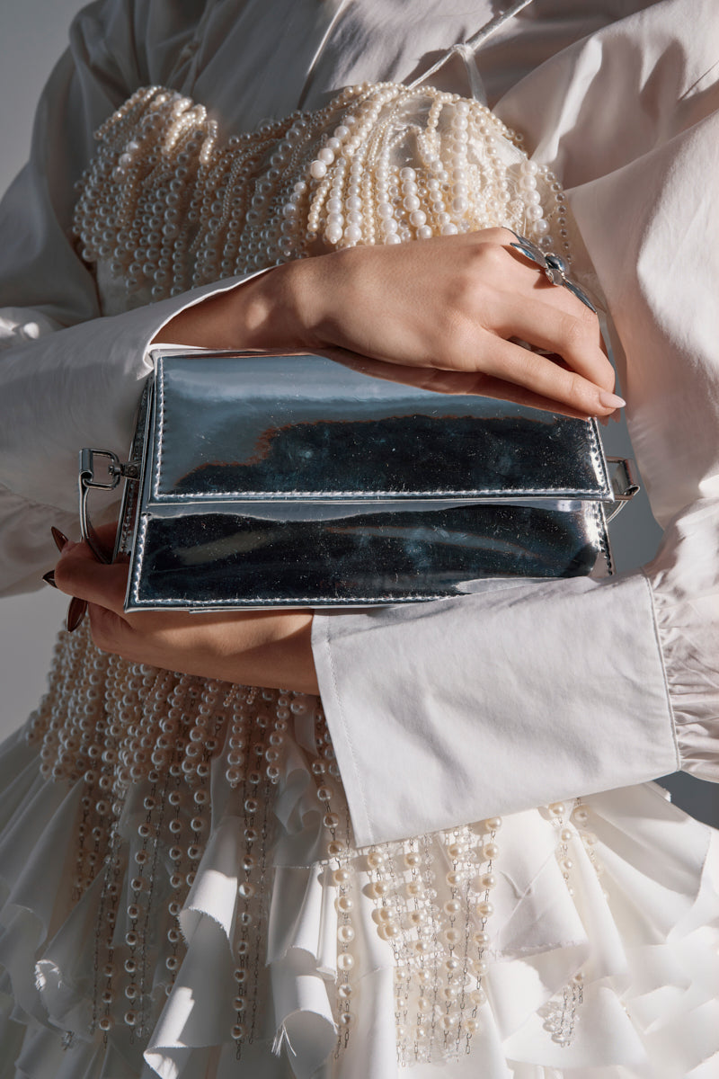 A person holds the I STAND ALONE CHROME PURSE close to their torso, wearing a white dress with strings of pearls and light pink nail polish.
