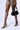A person is wearing AZALEA WANG JACINTO sandals with black spikes and a shiny, metal square-patterned skirt while holding a black handbag with silver accents. The background is a plain white studio setting.